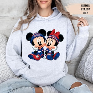 A woman wearing a grey hoodie with a graphic of two cartoon mice, one wearing a blue shirt and the other a red shirt, sitting together.