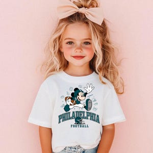 A young girl with blonde hair wearing a white t-shirt with a Philadelphia football graphic and a bow in her hair.