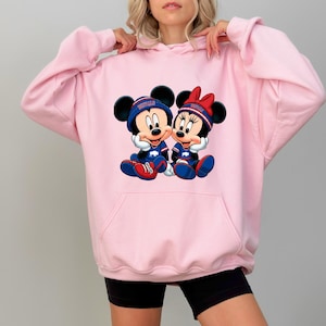 A woman wearing a pink hoodie with a graphic of two cartoon mice, Minnie Mouse and Mickey Mouse, sitting together.