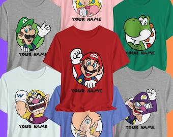 May include: Assortment of colorful t-shirts featuring cartoon characters from a popular video game series. Each shirt displays a different character in a circular design, with the text "YOUR NAME" below. The shirts are in various colors, including red, green, and gray.