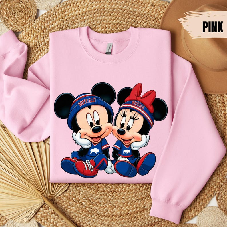 A pink sweatshirt with a cartoon image of Minnie Mouse and Mickey Mouse, along with the text "Buffalo" on their hats, is displayed on a woven mat.