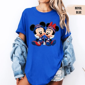 A woman wearing a blue t-shirt with a graphic of two cartoon mice, Minnie Mouse and Mickey Mouse, sitting together. She is also wearing a denim jacket and denim shorts.