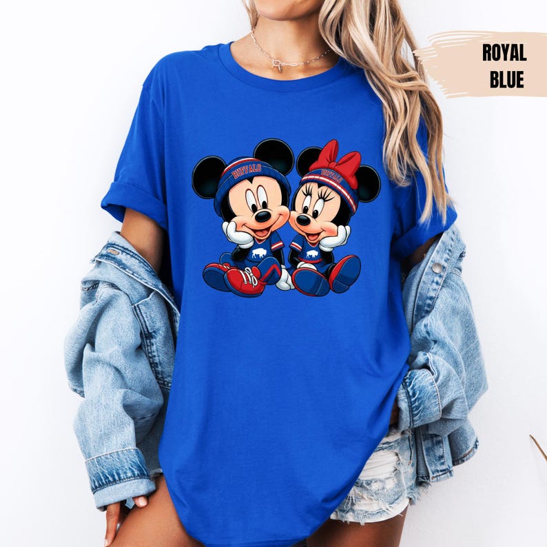 A woman wearing a blue t-shirt with a graphic of two cartoon mice, Minnie Mouse and Mickey Mouse, sitting together. She is also wearing a denim jacket and denim shorts.