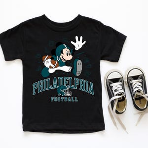 A black t-shirt with a graphic of a character from the Philadelphia Eagles football team, along with the team's logo and the text "PHILADELPHIA FOOTBALL". Next to the t-shirt is a pair of black and white sneakers.