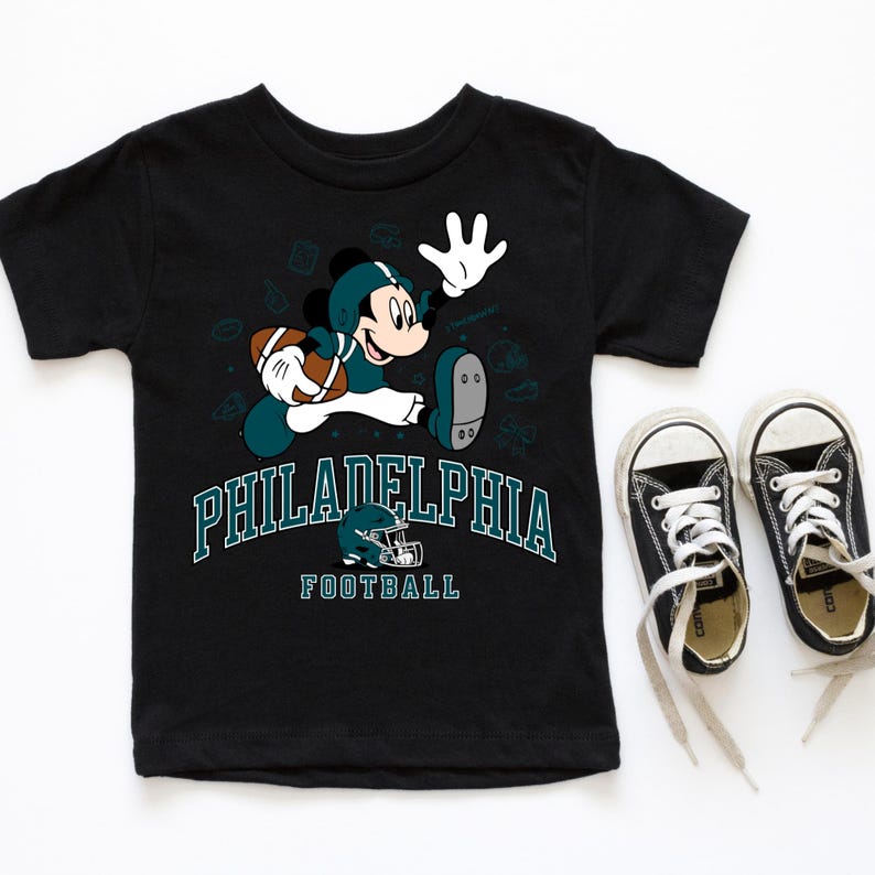 A black t-shirt with a graphic of a character from the Philadelphia Eagles football team, along with the team's logo and the text "PHILADELPHIA FOOTBALL". Next to the t-shirt is a pair of black and white sneakers.