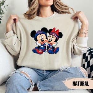 A woman wearing a beige sweater with a graphic of Minnie Mouse and Mickey Mouse, sitting on a couch.