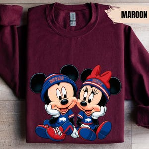A maroon t-shirt with a graphic design featuring two cartoon mice, one male and one female, wearing matching outfits and accessories.