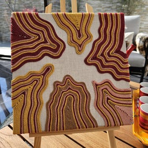 Puede incluir: Un lienzo cuadrado con un diseño abstracto colorido creado con hilo. El lienzo está montado en un caballete de madera.