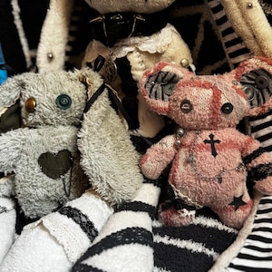 Puede incluir: Un conejo de peluche verde claro con un parche de corazón verde oscuro y un oso de peluche rosa con parches de estrellas negras, bordado de cruz y cadenas de metal. Una cinta métrica azul es visible a la izquierda, marcando 23 a 25 cm.
