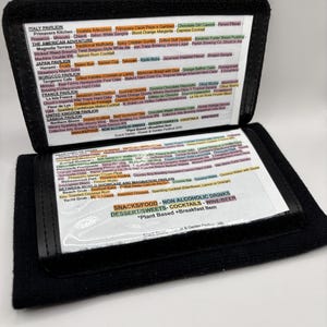 May include: A black leather menu holder with a clear plastic insert. The menu lists food and drink options from different pavilions at a festival. The pavilions include Italy, The American Adventure, Japan, Morocco, France, United Kingdom, and Canada. The menu also includes a section for snacks, food, non-alcoholic drinks, desserts, sweets, cocktails, wine, and beer.