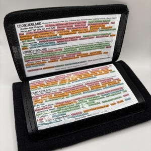 May include: A black leather wallet-style guide to Disney World restaurants, with a clear plastic window showing a list of restaurants and food items. The guide is open to the "Frontierland" and "Fantasyland" sections, with a list of restaurants and menu items in each section.