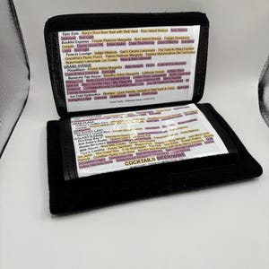 May include: A black leather wallet-style menu with a white background and pink text listing cocktails, beers, and wines. The menu is open to reveal a second page with more drinks listed. The text on the menu includes "Epic Eats", "Backlot Express", "Tune-in Lounge", "Grand Avenue", "PizzeRizzo", "BaseLine Tap House", "Toy Story Land", "Galaxy's Edge", "Docking Bay", "Kat Saka's Kettle", "Milk Stand", "Opa's Cantina", "Jet Juice", "Rancor", "Valley Harvest Brew", and "Cocktails Beer/Wine".