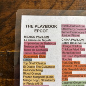 May include: A clear plastic menu from EPCOT's "The Playbook" with a list of food and drink items. The menu features sections for Mexico and China Pavilions, with items like "La Choza de Tequila" and "Orange Chicken". The text is in black font.