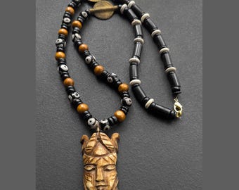 May include: A handcrafted necklace featuring a carved wooden mask pendant. The necklace has a beaded design with black, brown, and white beads. The mask pendant is light brown with carved facial features. The necklace is displayed on a dark gray surface.
