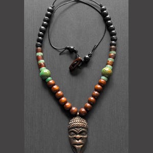 May include: A handcrafted necklace featuring a pendant of a tribal mask. The necklace has a black cord, wooden beads, and green and brown accent beads. The pendant is a silver color and has intricate details.