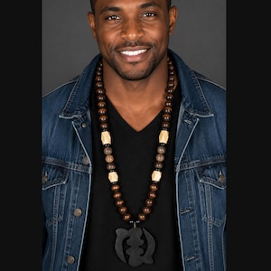 May include: A long beaded necklace featuring dark brown spherical beads, textured brown beads, and light-colored geometric wooden beads. The necklace includes a large, black, carved wooden pendant with a symmetrical, abstract, curved design.