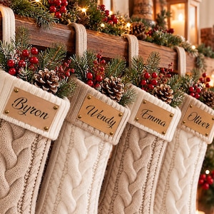 Puede incluir: Cuatro calcetines navideños de punto trenzado color crema colgados en fila. Cada calcetín tiene un parche rectangular de cuero tostado con los nombres Byron, Wendy, Emma y Oliver impresos en negro. Ramas de pino, piñas y bayas rojas.