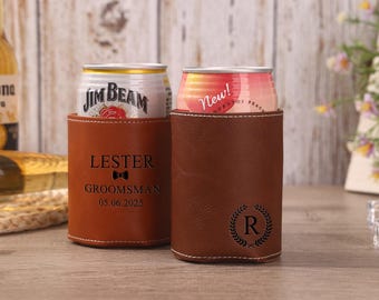 May include: Two brown leather can coolers. One is engraved with "LESTER GROOMSMAN 05.06.2025" and a bow tie. The other has a laurel wreath with the letter "R". Both hold cans.