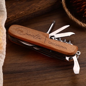 May include: A wooden multi-tool with various implements, including a knife, corkscrew, and screwdriver. The tool is engraved with the name "Daniella" and the word "Groomsman". The tool is on a dark wooden surface.