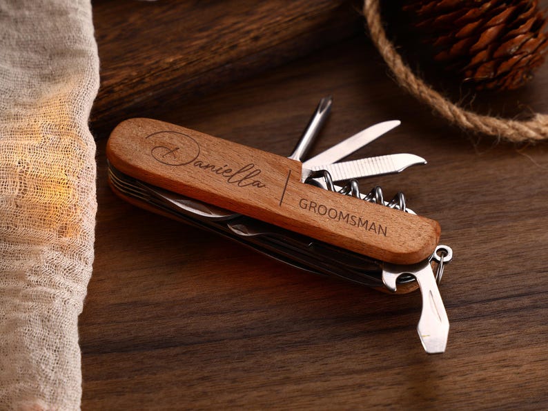 May include: A wooden multi-tool with various implements, including a knife, corkscrew, and screwdriver. The tool is engraved with the name "Daniella" and the word "Groomsman". The tool is on a dark wooden surface.