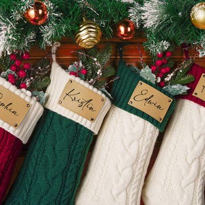 Puede incluir: Cuatro calcetines navideños de punto trenzado en rojo, verde y blanco, cada uno con una etiqueta de cuero personalizada. Decorados con vegetación y adornos festivos, perfectos para la decoración navideña.