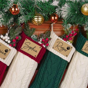 Może przedstawiać: Cztery świąteczne skarpety w warkoczowy wzór w kolorze czerwonym, białym i zielonym wiszą na drewnianym kominku, ozdobione świąteczną zielenią i ozdobami. Każda skarpeta posiada spersonalizowaną skórzaną metkę z imionami takimi jak "Janice" i "Roger".