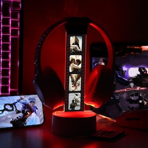 Peut inclure: Casque de jeu rouge sur un support éclairé avec une bande de photos. Un smartphone affiche une scène de jeu vidéo. Un clavier, une souris et une télécommande sont également visibles, tous éclairés en rouge, créant une configuration de jeu.