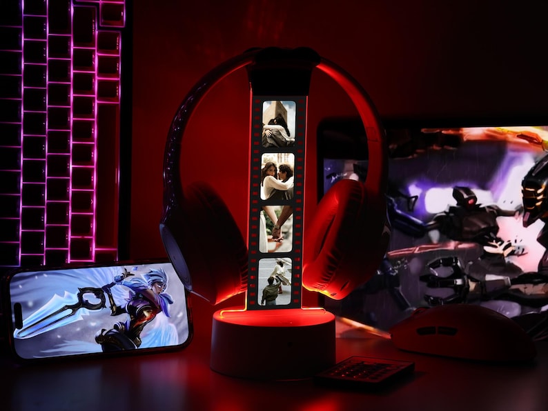 May include: Red gaming headphones on a lighted stand with a film strip of photos. A smartphone displays a video game scene. A keyboard, mouse, and remote are also visible, all illuminated with red light, creating a gaming setup.