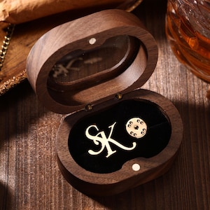 Op de afbeelding: Een open, ovaalvormige houten doos met een zwart fluwelen interieur. Binnenin zijn een goudkleurige speld en de letters "SK" zichtbaar. De doos staat op een houten oppervlak, met een sigaar en een glas amberkleurige vloeistof op de achtergrond.