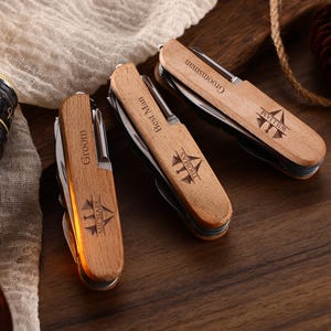 May include: Three personalized wooden pocket knives with silver blades are displayed on a wooden surface. Each knife is engraved with a name and title: "Groom Michael", "Best Man Thomas", and "Groomsman William". A bottle of liquor is visible in the background.
