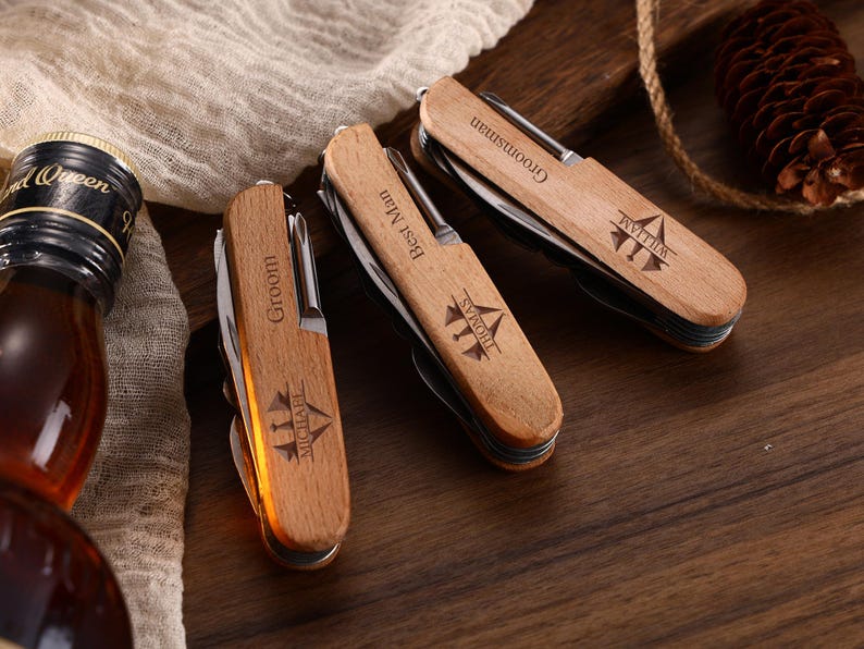 May include: Three personalized wooden pocket knives with silver blades are displayed on a wooden surface. Each knife is engraved with a name and title: "Groom Michael", "Best Man Thomas", and "Groomsman William". A bottle of liquor is visible in the background.