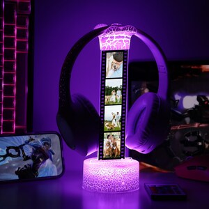 May include: A purple illuminated headphone stand with a film strip design displaying four photos. The stand has a cracked glass effect and supports a pair of purple headphones. A smartphone and keyboard are visible in the background.
