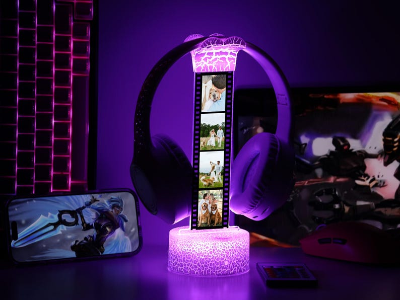 May include: A purple illuminated headphone stand with a film strip design displaying four photos. The stand has a cracked glass effect and supports a pair of purple headphones. A smartphone and keyboard are visible in the background.