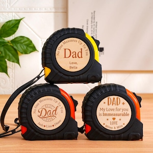 May include: Three black and yellow tape measures with personalized wooden faces. Each tape measure has the word "Dad" and a message. The tape measures are stacked on a wooden surface.