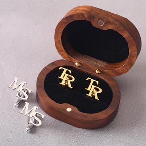 May include: Wooden oval box with gold cufflinks featuring the letters "TR" inside. The box is open, revealing the cufflinks on a black velvet lining. Two silver cufflinks with the letters "MS" are also visible.
