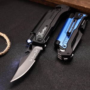 Puede incluir: Dos navajas de bolsillo, una negra y otra azul, sobre una superficie oscura. La navaja negra está abierta, revelando una hoja con el texto "We love you Dad". La navaja negra también tiene grabado el nombre "Thomas".