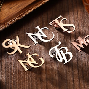 May include: A collection of gold and silver-toned initial pins arranged on a wooden surface. The pins feature various letter combinations, including "SK", "NC", "TS", "TB", "MP", and "NG". The pins are likely used as accessories or for personalization.