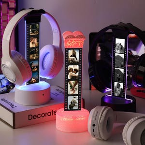 May include: Three illuminated headphone stands with personalized photo strips. One stand is heart-shaped, another is white, and the third is black. White and black headphones rest on the stands. The stands are on a white surface.