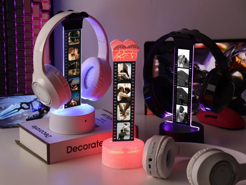 May include: Three illuminated headphone stands with personalized photo strips. One stand is heart-shaped, another is white, and the third is black. White and black headphones rest on the stands. The stands are on a white surface.