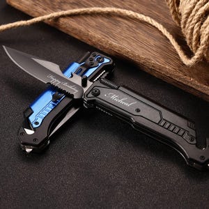 Könnte beinhalten: Ein schwarzes und blaues Klappmesser mit silberner Klinge. Die Klinge ist mit "Happy Birthday" graviert. Der Griff ist mit dem Namen "Michael" graviert. Das Messer liegt auf einer dunklen Oberfläche neben einem Holzbrett und einer Seilspule.