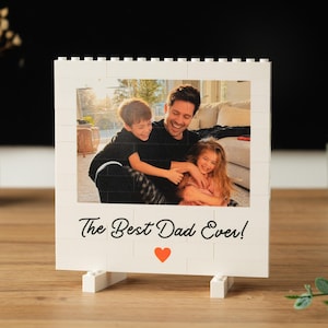 May include: A white brick-style photo frame displaying a family photo of a father and two children. The text "The Best Dad Ever!" is below the photo, with a red heart. The frame is on a wooden surface.