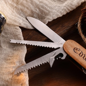 May include: Close-up of a multi-tool with a wooden handle engraved with the name "Edward". The tool has several open blades and a bottle opener. A bottle with a black and gold label is visible in the background.