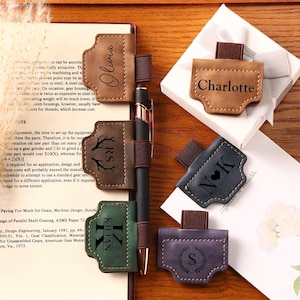 May include: A collection of personalized pen holders in various colors, including brown, green, blue, and purple. Each holder features a name or initial and a decorative design. A black pen is inserted in one of the holders.