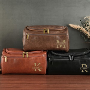 Könnte beinhalten: Drei personalisierte Kulturbeutel aus braunem, rotbraunem und schwarzem Leder. Jede Tasche hat einen goldenen Reißverschluss und einen Namen oder Initialen auf der Vorderseite. Die Taschen sind auf einer Holzoberfläche gestapelt.