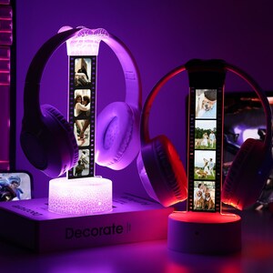 May include: Two illuminated headphone stands with personalized photo strips. One stand has purple headphones and a white base, the other has red headphones and a gray base. The stands are on a desk with a keyboard and monitor in the background.