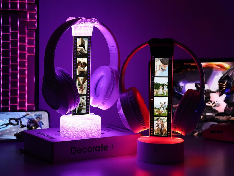 May include: Two illuminated headphone stands with personalized photo strips. One stand has purple headphones and a white base, the other has red headphones and a gray base. The stands are on a desk with a keyboard and monitor in the background.