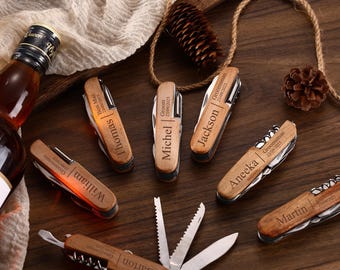 Puede incluir: Varios cuchillos de bolsillo de madera personalizados, grabados con nombres y fechas, se exhiben sobre una superficie de madera oscura. Una botella de licor y elementos decorativos completan la composición.
