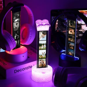 May include: A display of illuminated headphone stands with personalized photo strips. The stands feature a cylindrical base and a vertical photo strip, with a pair of headphones resting on top. The scene is bathed in purple and pink light.