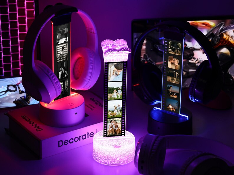 May include: A display of illuminated headphone stands with personalized photo strips. The stands feature a cylindrical base and a vertical photo strip, with a pair of headphones resting on top. The scene is bathed in purple and pink light.