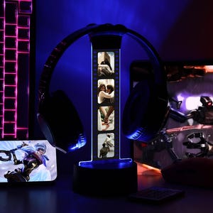 May include: A black headset rests on a lighted headphone stand displaying photos. A keyboard and a smartphone with a game graphic are visible. The scene is illuminated with blue and pink lights, creating a gaming setup atmosphere.